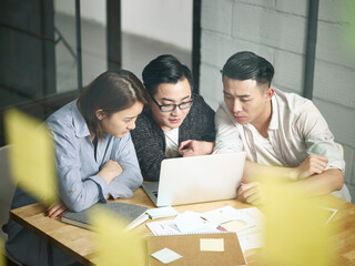 through the glass shot of a team of three young asian entrepreneurs discussing business in office using laptop computer