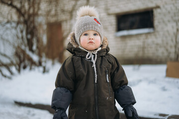 cute caucasian baby girl in winter overall and mittens standing in winter yard in Russia