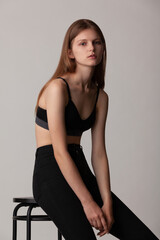 Naklejka premium beautiful girl in a black bra and black sweatpants posing while sitting on a high stool in the studio on a gray background