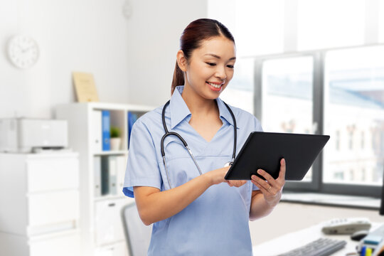 medicine, profession and healthcare concept - happy smiling asian female doctor or nurse in blue uniform with tablet pc computer and stethoscope over hospital background
