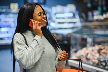 Fototapeta premium African american woman with shopping cart trolley in the supermarket store speak on mobile phone.