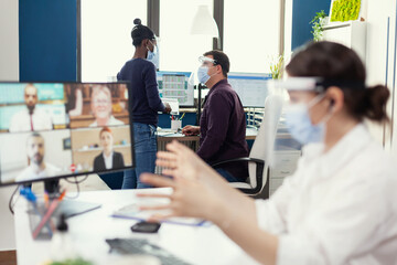 African woman having a conversation with her coworker waring face mask at workplace wearing face mask. Entrepreneur having videocall while colleagues working respecting social distance during global