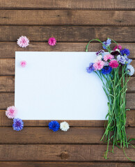 Colorful bouquet of summer garden flowers. Cornflowers with blank paper for greeting message on brown wooden table. Vintage floral background. Floral mock up with multicolored flowers. Copy space
