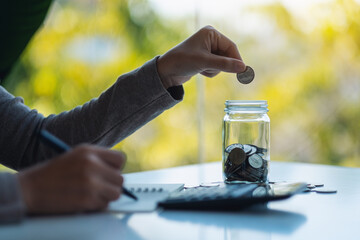 Closeup image of a woman putting coins in a glass jar , calculating and taking note for saving money and financial concept