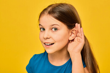 Hearing test concept. Positive girl holds hand near the ear, isolated on yellow background