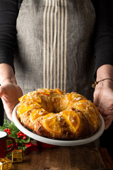 Woman with gray apron holding a white plate with freshly made Kings cake, on dark background,...