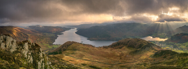 Naklejka na ściany i meble Panorama of dramatic clouds with a view over Ullswater in the Lake District as shafts of sunlight casts rays over Glenridding village