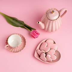 Creative layout made with teapot, cup, heart shaped cookies and tulip on pastel pink background. Festive table setting for Valentine's Day, Mother's Day or wedding. Minimal love concept.