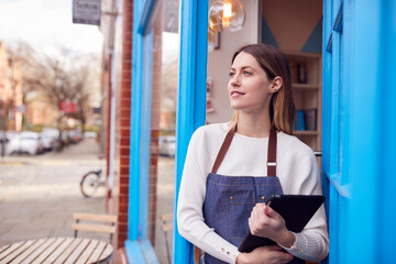 Female Small Business Owner With Digital Tablet Standing In Shop Doorway On Local High Street