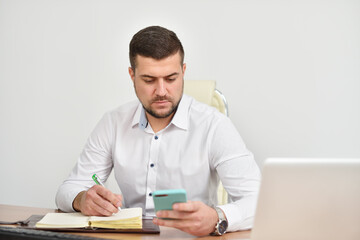 Obraz premium Młody kierownik w biurze uważnie przygląda się smartfonowi i zapisuje numer w notesie