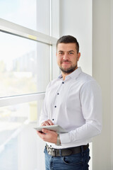Fototapeta premium Portrait of a young male manager in the office with a tablet computer stands by a large window