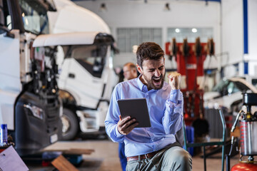 Young bearded excited chief holding tablet and cheering for success. Garage of shipping firm interior. In background are trucks.