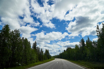 Naklejka premium View from relief car windscreen on the blue sky with white clouds, grey asphalt road and landscape with forest and green teeses. Landscape through window