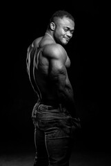 Black fitness model man stands half turned to the camera on black background. Naked torso of very muscular man.