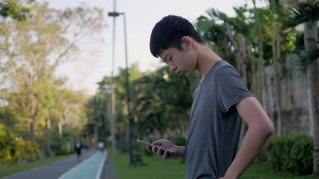 A tall, thin, black-haired young Asian man playing smartphone at the park. Expressing joy, happiness, and fulfillment. A young man plays a cell phone to check information on social media.