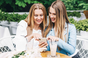 A beautiful girl spends time with her mother and shows her something interesting in her phone. Two beautiful blonde women on a summer terrace