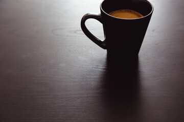 Black cup of hot coffee on dark wooden background, back lit