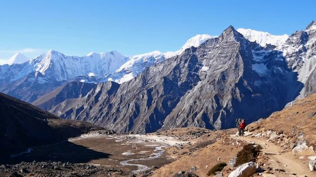 Trekker and porter with backpacks on the western ascent to Renjo La Pass in Everest region in the Nepali Himalayas with valley and rugged snow-covered mountains on sunny day on Three Passes Trek.
