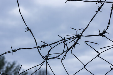 Metal barbed wire against an overcast blue sky in spring or winter and a hole in the fence. Protecting a parking lot, business or jail. 
