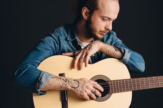 Tattooed musician posing with guitar over black background. Stylish guy with acoustic guitar working in studio