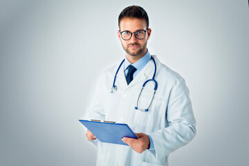 Studio portrait shot of young male doctor holding clipboard in his hand while standing at isolated...