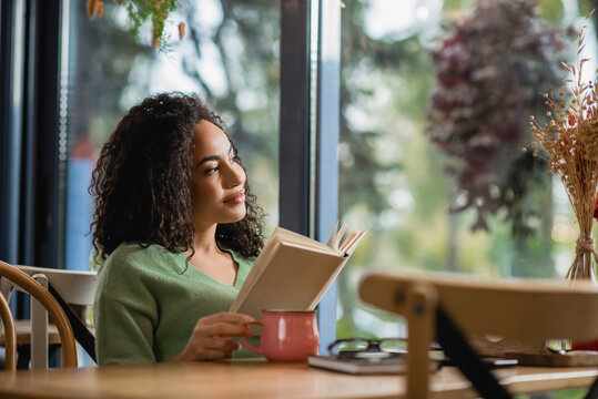 Dreamy African American Woman Holding Book And Looking At Window In Cafe