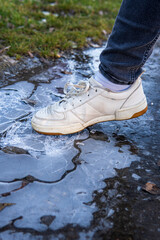 A woman in white sneakers breaking or crushing ice on a pathway in a park. Cracked ice under woman's feet on a  cold, winter, day. 
