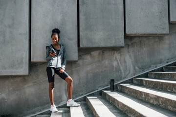 Fototapeta premium Uses phone. Young african american woman in sportive clothes have workout outdoors at daytime