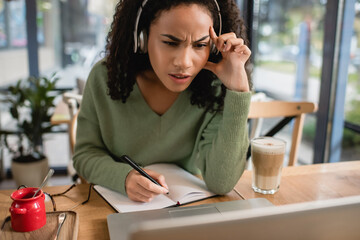 focused african american woman listening podcast and looking at laptop in cafe