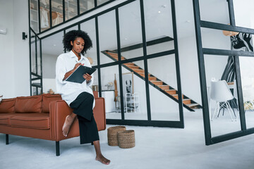 Holds notepad. Young african american woman with curly hair indoors at home