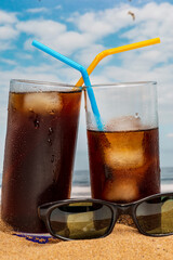 Two glasses of soda with ice and straws on the beach with sunglasses and a blue sky with seagulls flying in Tenerife, Canary Islands.