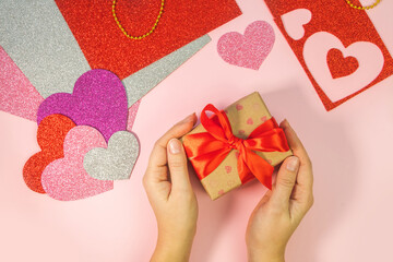 Packaging Valentine's Day or birthday presents. Valentine's Day gifts with a red paper heart on a pink background. Top view. Female hands hold gift box with red ribbon.