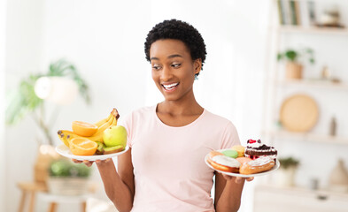 Cheerful black woman holding plates with fruits and desserts indoors, choosing between healthy and...