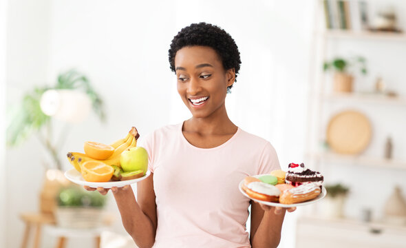 Cheerful black woman holding plates with fruits and desserts indoors, choosing between healthy and junk foods