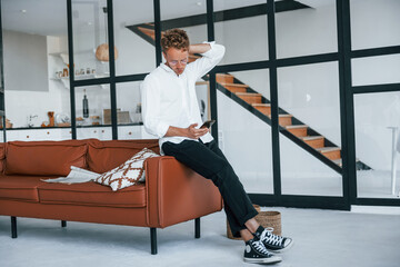 Holds phone in hand. Caucasian young guy in elegant white shirt indoors at home