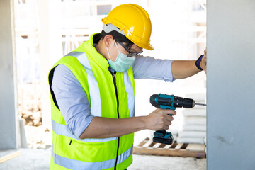 Construction foreman worker in hardhat safety helmet drilling by electric drill on the construction site. wearing surgical face mask during coronavirus covid and flu outbreak