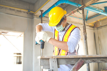 Construction foreman worker in hardhat safety helmet drilling by electric drill on the construction site. wearing surgical face mask during coronavirus covid and flu outbreak