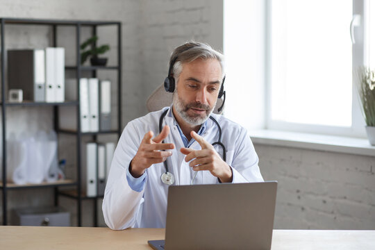 Portrait of senior grey-haired male doctor in his office using laptop for video chat with a patient. Online consultation with doctor for diagnoses and treatment recommendation. Telehealth concept.