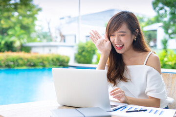 An Asian female office worker taking video calls, online conferences, smiling and greeting colleagues through her laptop at the poolside table, the concept of a home-made video call via the Internet.