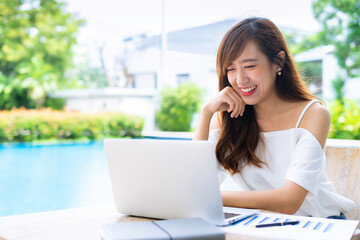 An Asian female office worker taking video calls, online conferences, smiling and greeting colleagues through her laptop at the poolside table, the concept of a home-made video call via the Internet.