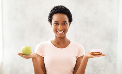 Choice between wholesome and unhealthy food. Happy black woman holding apple and donut, choosing...