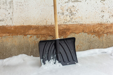 A black shovel with a wooden handle for snow removal stands against the old wall.