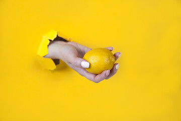 A woman's hand passes a lemon through a hole in a yellow paper background. Health concept, wholesome food.