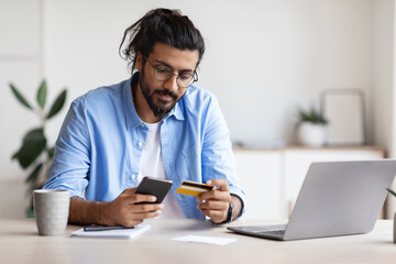 Arab freelancer guy using smartphone and credit card at home, paying online