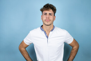 Young handsome man wearing a white t-shirt over blue background skeptic and nervous, disapproving expression on face with arms in waist