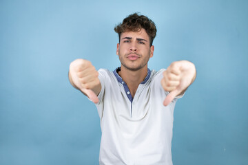 Naklejka premium Young handsome man wearing a white t-shirt over blue background with angry face, negative sign showing dislike with thumb down