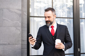 Selective focus at face. Adult Caucasian business man with beard, dress in formal suit standing in central business district. While drink morning coffee and look  at his mobile smartphone.