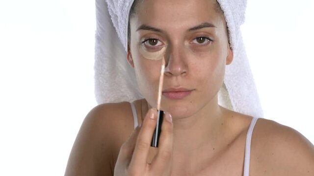 A young woman applies concealer under her eyes and smears it with her fingers on a white background
