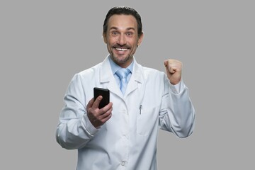Joyful scientist holding cell phone and gesturing success. Happy caucasian doctor with mobile phone showing victory gesture against gray background.
