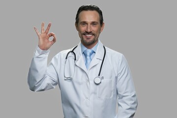 Portrait of handsome doctor showing ok sign. Cheerful male practitioner with stethoscope looking at camera on gray background.
